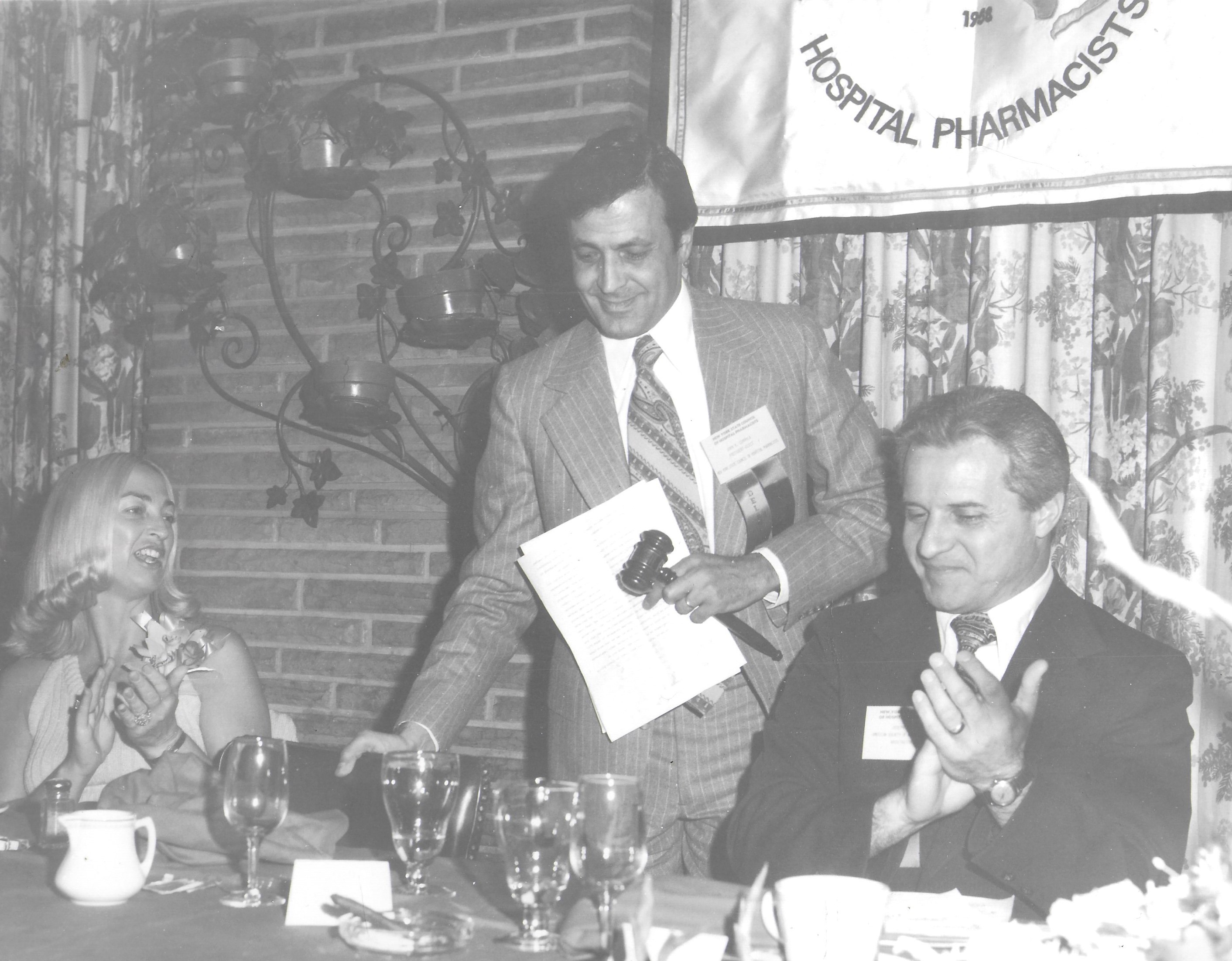 A black and white photo of a dinner in 1974. A man with a gavel and a name badge stands between a woman and man at a table, both sitting