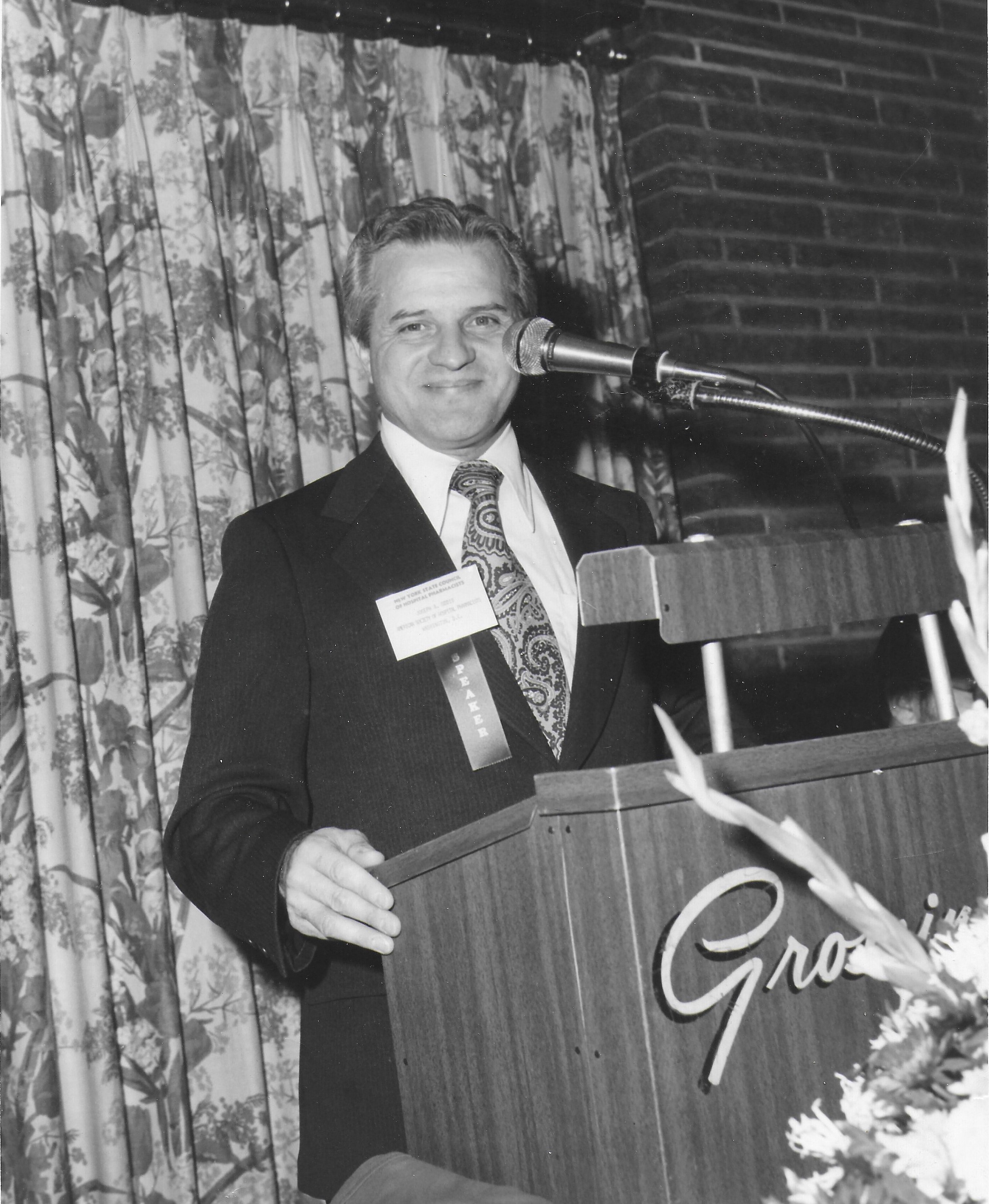 A smiling man in a suit and name badge stands at a podium